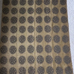 Puede incluir: Tela dorada con un patrón repetitivo de diseños circulares de color gris oscuro. Cada círculo tiene un centro punteado y un borde exterior puntiagudo, creando una apariencia texturizada. La tela tiene un brillo sutil.