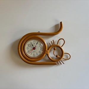 May include: A decorative wall clock with a unique design. The clock features a round face with black numerals and red hands, encased in a light brown, woven rattan frame. The frame extends into a whimsical, stylized shape resembling a cat or mouse.