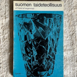May include: A book titled "Suomen taideteollisuus" with a blue cover featuring a black, abstract glass design. The author's name, Ulf H&aring;rd af Segerstad, is also visible. The publisher's name, Tammi, is at the bottom.