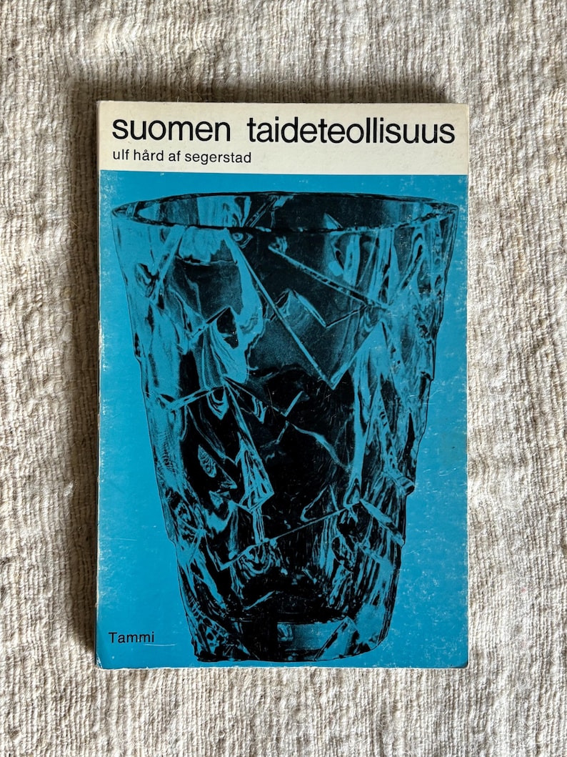 May include: A book titled "Suomen taideteollisuus" with a blue cover featuring a black, abstract glass design. The author's name, Ulf H&aring;rd af Segerstad, is also visible. The publisher's name, Tammi, is at the bottom.