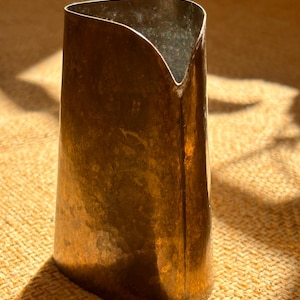 Op de afbeelding: Een hoge, bronskleurige metalen vaas met een unieke, asymmetrische bovenkant. De vaas heeft een taps toelopende vorm, breder aan de basis en smaller naar de bovenkant toe. Het oppervlak heeft een textuur en de belichting creëert schaduwen.