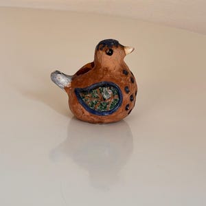 May include: A small, brown ceramic bird figurine with blue and green painted details. The bird has a white beak and a blue and white tail.