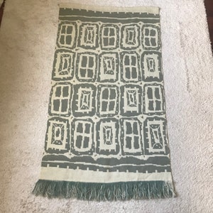 Puede incluir: Alfombra rectangular con un patrón repetitivo de rectángulos grises sobre un fondo crema. La alfombra tiene flecos en el borde inferior. El diseño es geométrico y moderno, adecuado para una sala de estar o dormitorio.