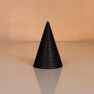 以下が含まれることがあります： 黒色の円錐形オブジェクト。表面にはテクスチャがあり、直立しています。下の反射面に三角形の影を落としています。背景は単色の明るいピーチ色です。