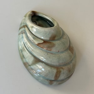 Könnte beinhalten: Ein Keramik-Wandtopf in Form einer Muschel mit hellgrüner Glasur und braunen und weißen Marmorierungen. Der Topf hat oben ein Loch zum Aufhängen.