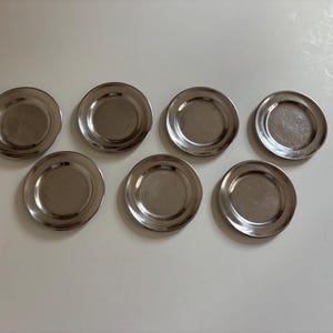 May include: Seven small, round, silver-colored metal plates are arranged on a white surface. The plates have a slightly concave center and a raised rim. They appear to be coasters or small serving dishes.