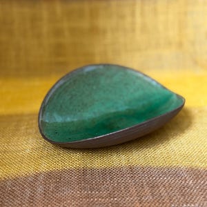 Peut inclure: Un plat en céramique en forme de goutte d'eau avec un intérieur vert émeraude brillant et un bord marron foncé. Le plat est posé sur un fond rayé jaune et marron, soulignant sa forme et sa couleur uniques.