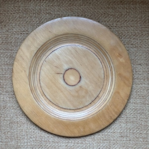 May include: A circular wooden plate in a light colour, featuring concentric grooves and a central circle. The plate has a natural wood grain and a simple, rustic design. It is likely used for serving or display.