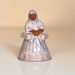 Può includere: Statuetta in ceramica di una donna con un mantello bianco con cappuccio e una gonna a righe. La statuetta ha un cappuccio azzurro e strisce verticali blu sulla gonna. La base ha il testo "KRINGEL-STAVA".