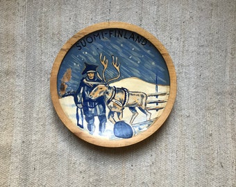 May include: A round wooden plate with a painted scene of a person in a blue coat and hat with a reindeer pulling a sled. The plate has the text "SUOMI-FINLAND" around the edge.
