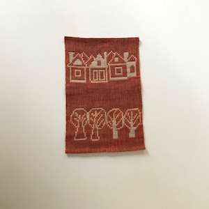 Puede incluir: Un tapiz tejido para colgar en la pared con fondo marrón y casas y árboles blancos. Las casas tienen techos y ventanas blancas. Los árboles tienen troncos y ramas blancas.