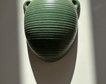 May include: A green ceramic wall vase with a ribbed texture and two small handles. The vase is oval-shaped and casts a long shadow on a white surface. The vase is a decorative item for home decor.