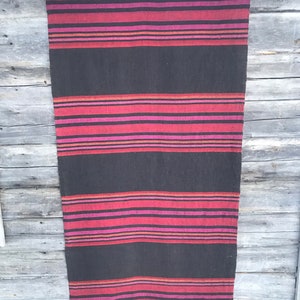 Puede incluir: Una alfombra tejida a rayas negras y rojas colgada de una pared de madera. La alfombra tiene un diseño tradicional con rayas alternas de negro y rojo, con algunas rayas finas de rosa y naranja.