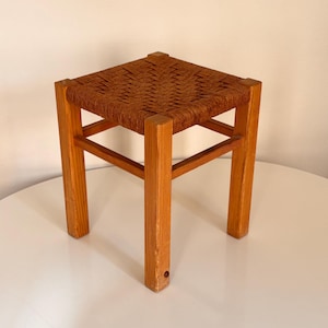 May include: A small, square wooden stool with a woven seat. The frame is made of light-colored wood, and the seat is a warm brown color. The stool has four legs and is designed for sitting.