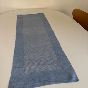 Può includere: Una tovaglia blu rettangolare con un centro blu più chiaro. La tovaglia è realizzata in tessuto intrecciato e viene esposta su un tavolo bianco. I bordi della tovaglia sono di una tonalità di blu più scura.