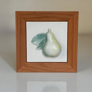 以下が含まれることがあります： 梨と葉が描かれた額入りのセラミックタイル。梨は薄緑色で、背景は白です。フレームは木製です。このアートワークは装飾品です。