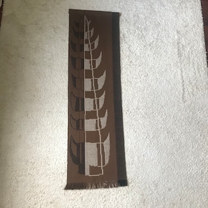 Puede incluir: Una bufanda marrón y gris con un diseño geométrico. La bufanda presenta un patrón repetitivo de formas estilizadas y un borde con flecos. La bufanda es rectangular y parece estar hecha de tela tejida.