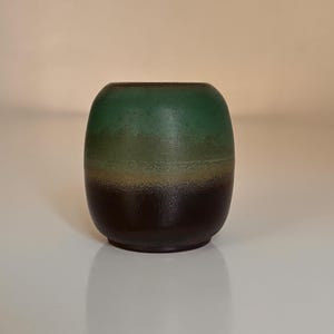 May include: A small, rounded ceramic vase with a gradient of colours. The top is a deep green, transitioning to a lighter green, then to a tan, and finally to a dark brown base. The vase has a smooth, matte finish.