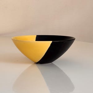 May include: A ceramic bowl with a striking design. The bowl is divided into two halves: one in a vibrant yellow and the other in a deep black. The bowl's shape is a simple, yet elegant, cone. The bowl is likely used for serving food or as a decorative item.