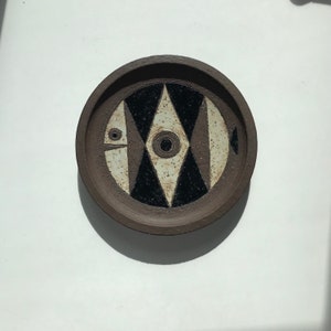 以下が含まれることがあります： 白と黒の幾何学模様が施された茶色の陶器の皿。魚の形をしていて、大きな目とスタイリッシュな体が特徴です。