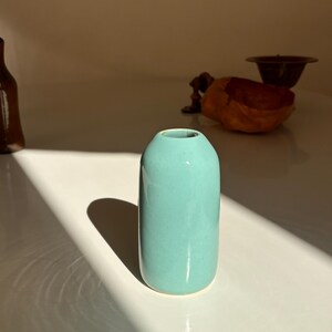 Könnte beinhalten: Eine kleine, zylindrische, helltürkisfarbene Keramikvase. Die Vase hat eine abgerundete Oberseite und eine glatte, glänzende Oberfläche. Die Vase steht auf einer weißen Oberfläche, mit einem Schatten nach links.