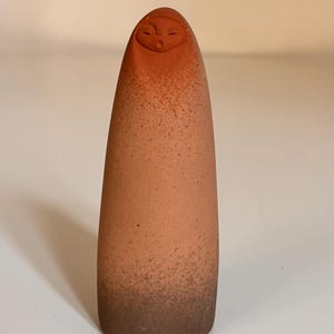Op de afbeelding: Een terracotta-kleurige, langwerpige figuur met een afgeronde bovenkant en een gezicht dat in het bovenste gedeelte is gesneden. Het beeldje heeft een gespikkelde textuur en een kleurverloop, dat overgaat van een lichter oranjebruin aan de bovenkant naar een donkerder bruin aan de basis.
