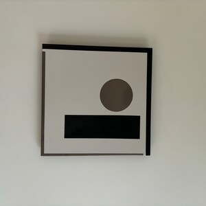 以下が含まれることがあります： 黒いフレームの抽象的な幾何学的なアートプリント。アートワークは、白い背景に灰色の円と黒い長方形を含んでいます。この作品は白い壁に取り付けられています。フレームは正方形です。
