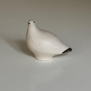 May include: A ceramic dove figurine in a matte white finish. The bird has a rounded body, small black eyes, and a dark gray tail. The dove is a symbol of peace and is often used as a decorative item.