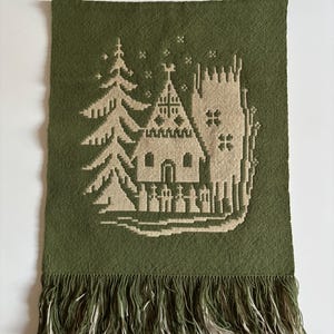 Puede incluir: Tapiz de pared tejido verde con un diseño de iglesia y árbol beige. El tapiz rectangular tiene un borde inferior con flecos. La iglesia presenta un campanario y una cruz, con un árbol a la izquierda. El fondo es verde oliva liso.