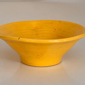 Puede incluir: Un cuenco de cerámica amarillo brillante con forma acampanada. El cuenco tiene un borde ancho y una superficie ligeramente texturizada. El cuenco es probablemente hecho a mano y adecuado para servir alimentos o como pieza decorativa.