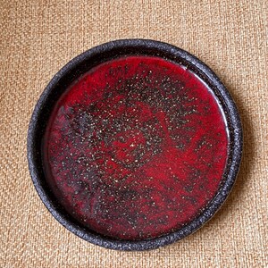 以下が含まれることがあります： 深紅色の内側と黒いテクスチャの縁を持つ丸いセラミックプレート。赤い表面には小さな暗い粒子が散りばめられており、独特の視覚的テクスチャを作り出しています。このプレートはおそらく食べ物の提供に使用されます。