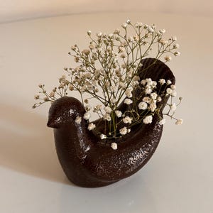 May include: A dark brown, bird-shaped ceramic vase with a glossy, textured finish. The hollow back of the bird holds a cluster of delicate, small white baby's breath flowers that extend upwards and outwards from the center of the vessel.