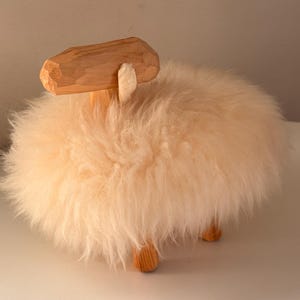 Könnte beinhalten: Eine handgefertigte Holzschaf-Figur mit einem flauschigen, cremefarbenen Wollkörper. Das Schaf hat einen geschnitzten Holzkopf und Beine, was eine charmante, rustikale Ästhetik erzeugt. Die weiche Wolle bildet einen taktilen Kontrast zum glatten Holz.