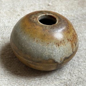 以下が含まれることがあります： 茶色、白、黄色の斑点のある釉薬が施された、丸い小さな陶器の花瓶。花瓶の上部に広い開口部があります。