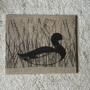 Könnte beinhalten: Eine schwarze Silhouette einer Ente, die in einem Feld mit hohem Gras schwimmt, gedruckt auf einem natürlichen Leinenstoff.