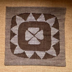 Puede incluir: Textil cuadrado tejido con un diseño geométrico. El cuadrado central es marrón oscuro, con un patrón floral gris-azulado más claro en el centro. El borde es de un marrón grisáceo más claro y el fondo es de color beige.