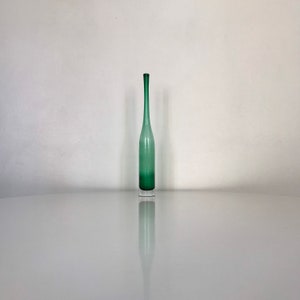 Puede incluir: Un jarrón de vidrio verde, alto y delgado, con un cuello largo y una base transparente. El jarrón tiene un degradado de color, que va de un verde más oscuro en la base a un verde más claro hacia la parte superior. El jarrón se exhibe sobre una superficie reflectante.