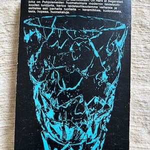 May include: A book cover featuring a black background with a turquoise geometric design of a vase. The cover includes text in Finnish, with the number 76 at the bottom right and the number 5 in parentheses at the bottom left.