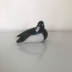 Op de afbeelding: Een zwart-witte keramische eendenfiguur. De eend ligt op zijn zij met zijn kop naar de zijkant gedraaid. De eend heeft een zwarte kop en lichaam met een witte buik.