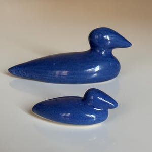 Puede incluir: Dos figuras de patos de cerámica en un tono azul profundo brillante. El pato más grande es alargado, mientras que el más pequeño es más compacto. Ambos tienen una forma suave y redondeada, lo que sugiere un diseño minimalista. Artículos decorativos.