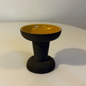 以下が含まれることがあります： 黒いセラミック製の台座に、黄色のボウルが載っています。台座は丸みを帯びたベースと、背の高い円筒形のステムを持っています。