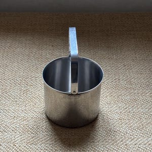 Pode incluir: Um cortador de biscoitos em aço inoxidável com corpo cilíndrico e uma pega retangular. A pega está presa no topo do cortador. O cortador está sobre uma superfície texturizada bege. Diâmetro de aproximadamente 7 cm.