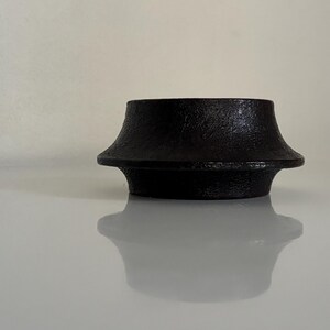 May include: A black, cast iron object with a textured surface. The object has a flared top and a wide base, with a slight hourglass shape. The object is sitting on a reflective surface.