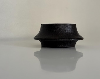 Op de afbeelding: Een zwart object van gietijzer met een getextureerd oppervlak. Het object heeft een uitlopende bovenkant en een brede basis, met een lichte zandlopervorm. Het object staat op een reflecterend oppervlak.