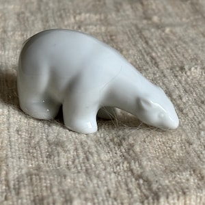 Puede incluir: Pequeña figura de oso polar de cerámica blanca. El oso está representado en posición de caminar, con la cabeza baja. La figura tiene superficies lisas y brillantes y descansa sobre una superficie texturizada de color claro.