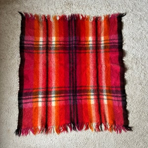 May include: A square, fringed blanket with a plaid pattern in shades of red, orange, and black. The blanket has a soft, textured appearance and is likely made of wool or a similar material. The fringe is along the edges.