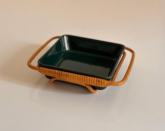 May include: A dark green square ceramic dish is presented in a woven rattan holder with two curved handles. The dish and holder are set against a neutral background, highlighting the contrasting textures and colours of the materials.