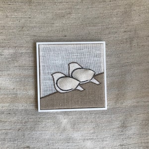 Puede incluir: Una pieza de bordado enmarcada con dos pájaros blancos con contornos marrones, posados sobre un fondo marrón. Los pájaros están mirando en la misma dirección, con el pájaro de atrás superponiéndose ligeramente al pájaro de delante.