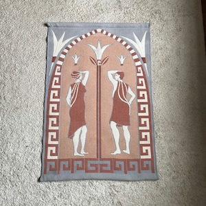 Puede incluir: Un tapiz rectangular con un diseño clásico. Dos figuras con túnicas de color terracota se encuentran a ambos lados de una columna central. El fondo es melocotón, enmarcado por un borde gris con un patrón de clave griega y detalles blancos.