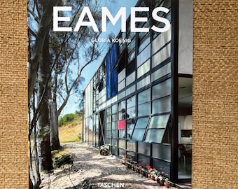 Peut inclure: Livre intitulé "EAMES" de Gloria Koenig, publié par Taschen. La couverture présente une photographie d'un bâtiment moderne avec de grandes fenêtres et un accent bleu. Le livre est posé sur un fond texturé et beige.
